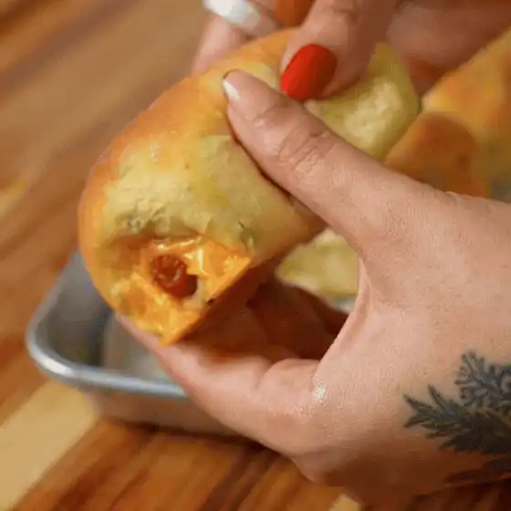 A person holds a baked bread roll filled with melted cheese and pieces of sausage over a metal baking tray. - Hungry Mockingbird