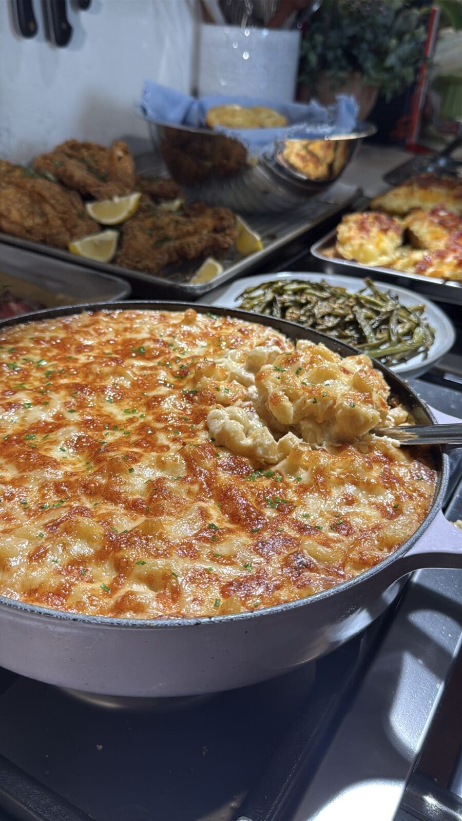 A large skillet of baked white cheddar mac and cheese with a portion being scooped, with side dishes including breaded cutlets and green beans in the background. - Hungry Mockingbird