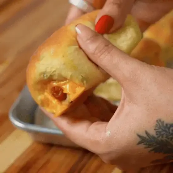 A person holds a baked bread roll filled with melted cheese and pieces of sausage over a metal baking tray. - Hungry Mockingbird