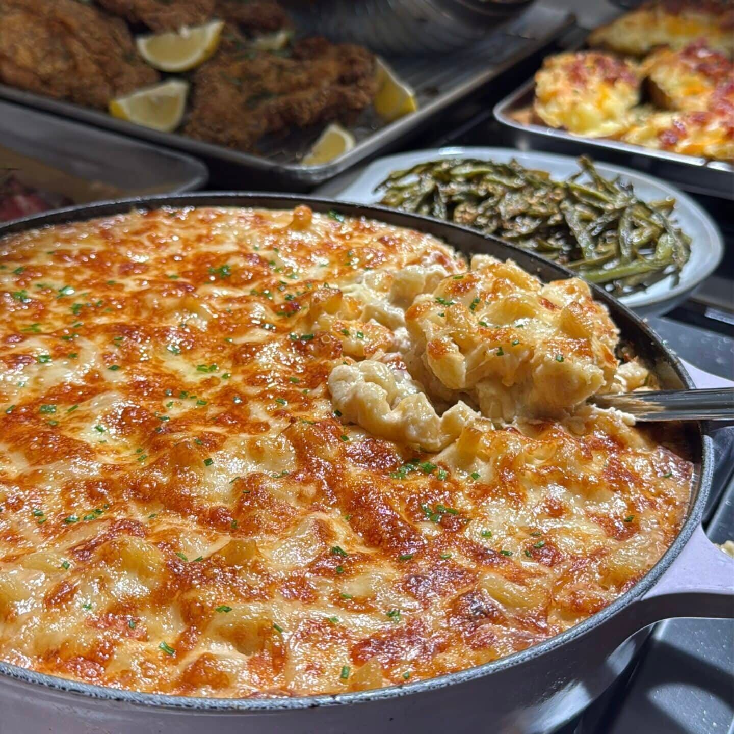 A large skillet of baked white cheddar mac and cheese with a portion being scooped, with side dishes including breaded cutlets and green beans in the background. - Hungry Mockingbird
