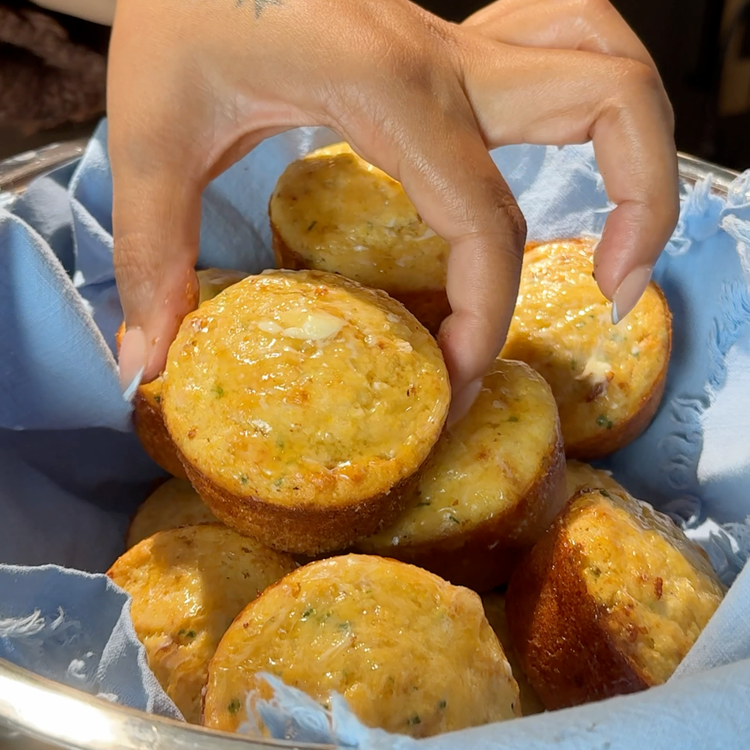 A hand picks up a homemade cornbread muffin from a basket lined with a blue cloth, filled with several golden-brown savory muffins. - Hungry Mockingbird