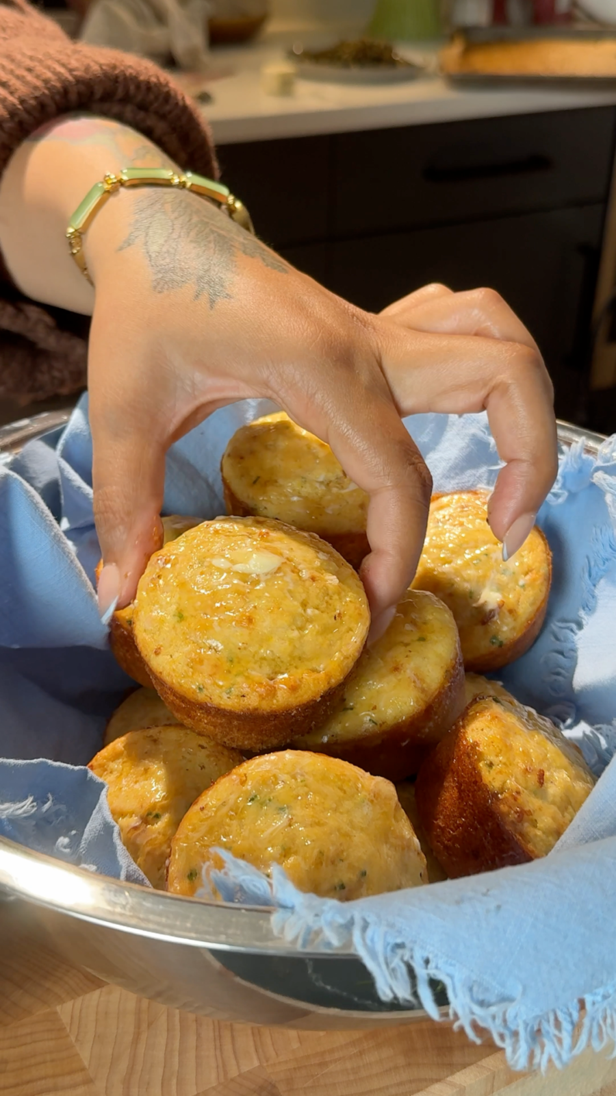 A hand picks up a homemade cornbread muffin from a basket lined with a blue cloth, filled with several golden-brown savory muffins. - Hungry Mockingbird